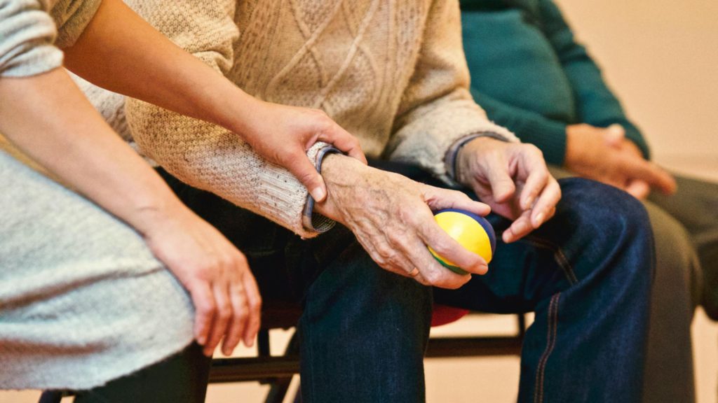 An older person's hand holding a ball. Sitting next to them is someone who has a friendly hand on the older person's wrist.